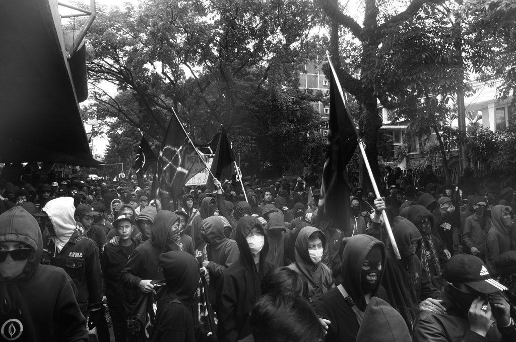 A crowd of people marching in a street. Most of them are dressed in black and masked. Several black flags can be seen in the crowd.