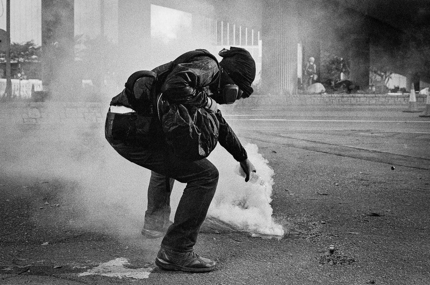 In a street, a person fully dressed in black and masked is pouring water on an object that is emitting a white smoke resembling tear gas. Their jacket is hiking slightly up, revealing the color of either their skin or the layer of clothing underneath their black clothing.