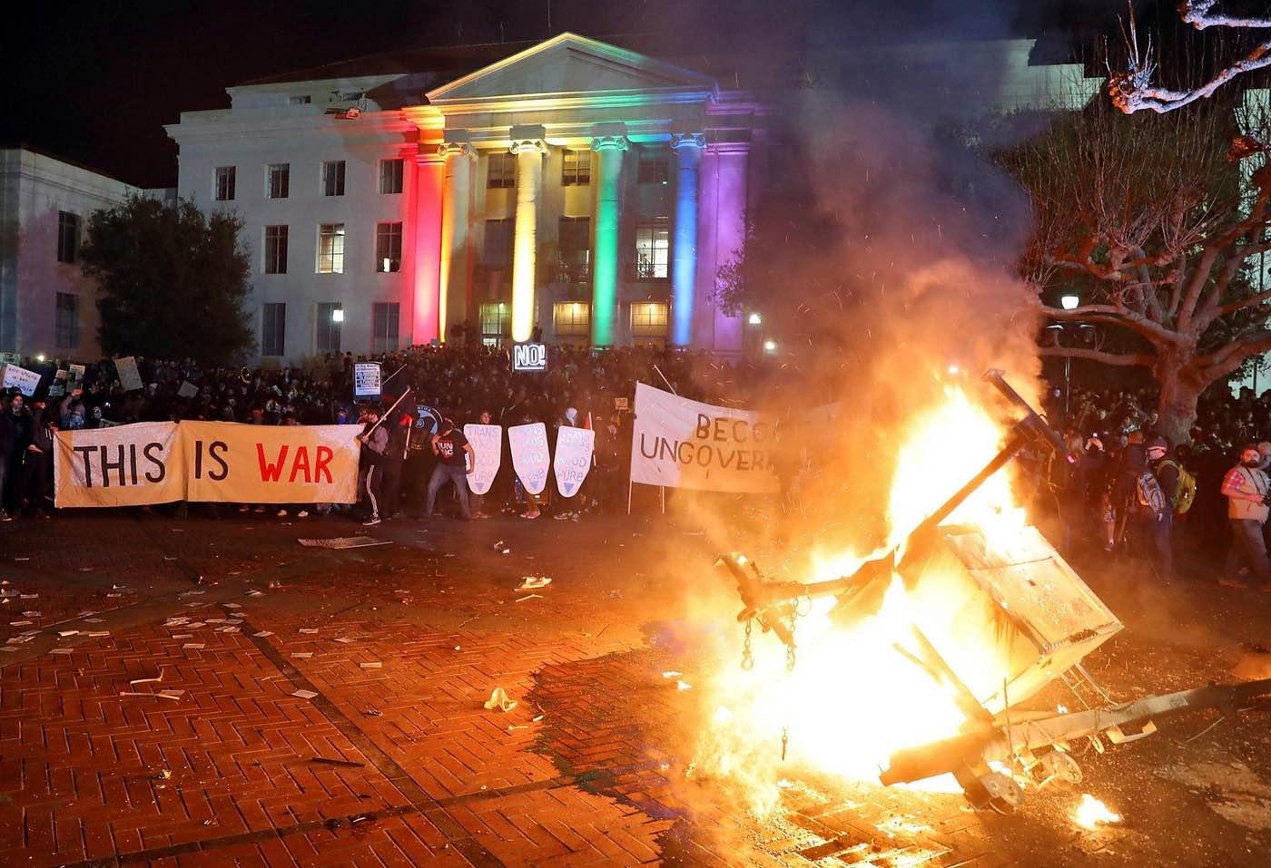 A crowd stands in the street at night. Behind the crowd is a large building. In front of the crowd, on the pavement is a small fire. Some people in the crowd are holding a banner reading "THIS IS WAR". Others are holding makeshift shields.