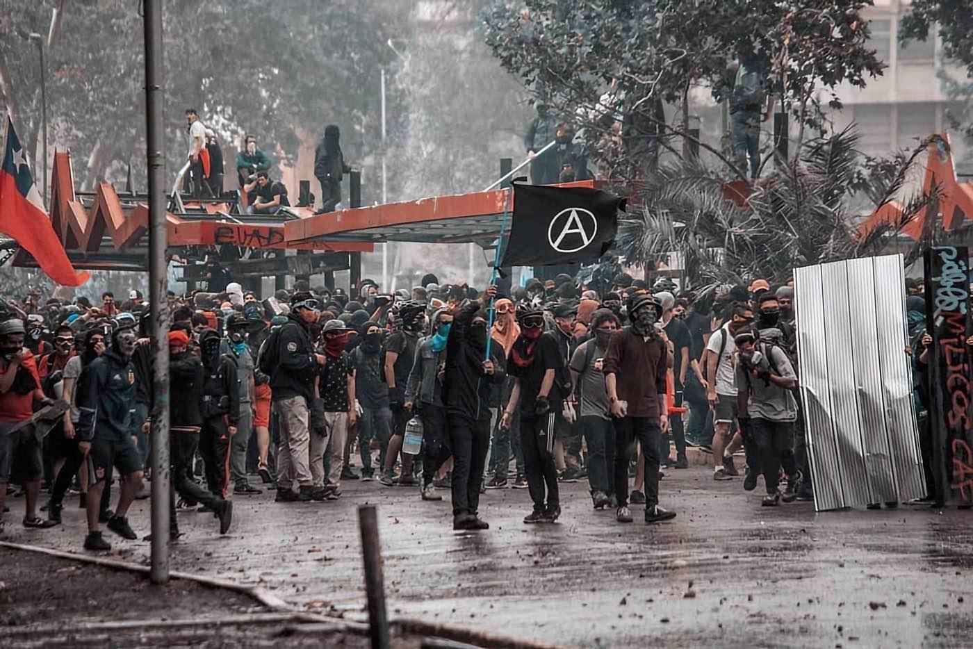 A crowd stands in a street, some of them are dressed in black and some of them wear protective equipment such as goggles and gas masks. A black flag with a "circled A" symbol is visible in the crowd.