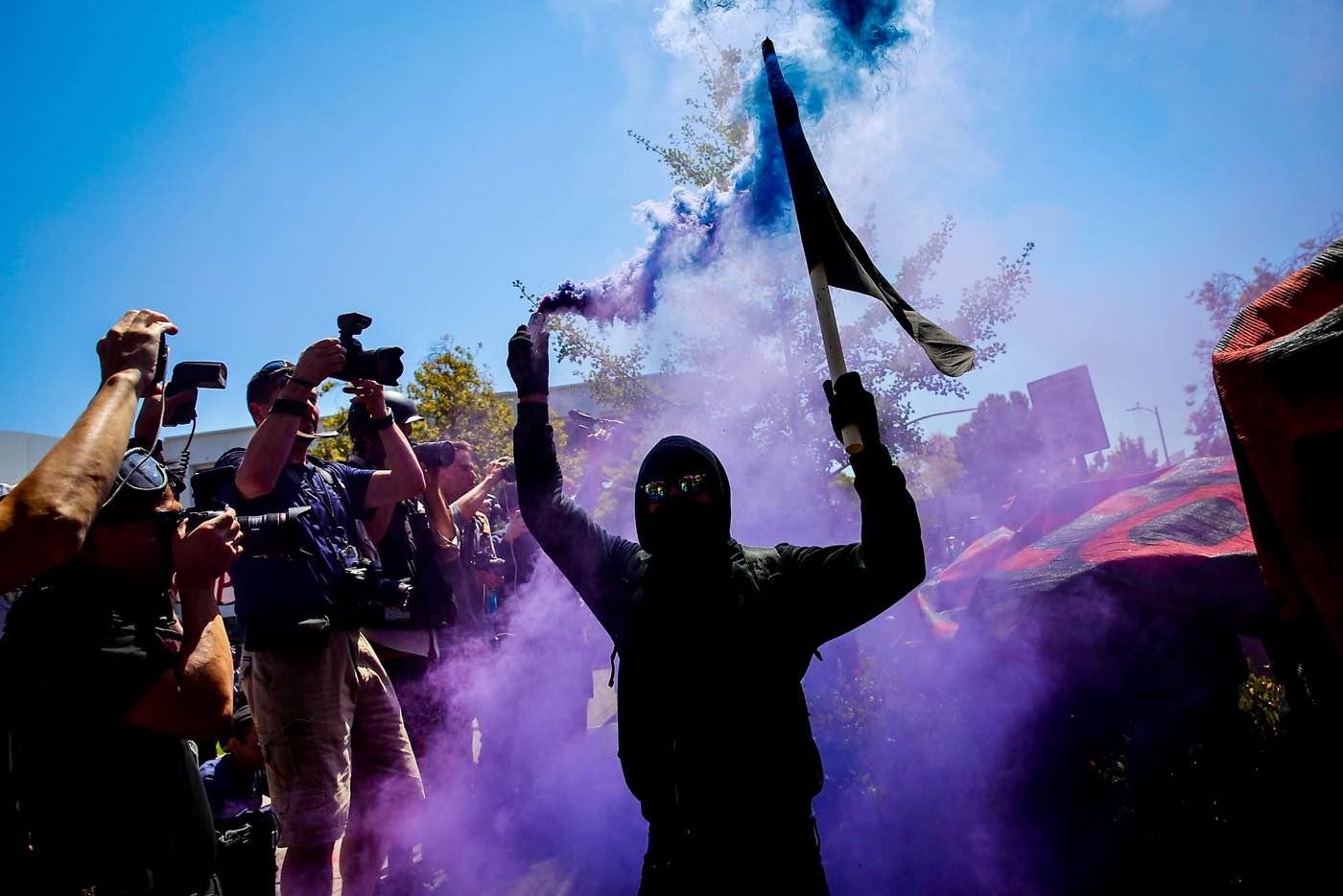 A person fully dressed in black is holding a black flag in one hand and a lit blue smoke grenade in the other. On one side of the person there seems to be an ongoing demonstration. On the other side there are many photographers with their cameras pointed at the demonstration.