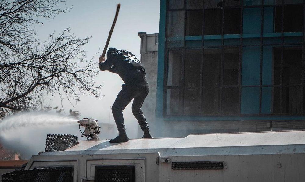 A person dressed in black is standing on top of a water cannon that is shooting water. In front of them is the cannon's device that is shooting water. They are holding a stick as if they are about to strike the device with it. They seem to be fully dressed in black except that they are not wearing gloves and that their top layer of clothing features a white Chaos Star.